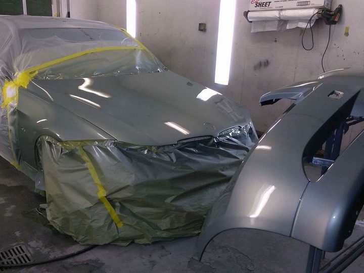 A car covered in masking film and yellow tape in the paint area, with a silver bumper standing on the floor beside it.